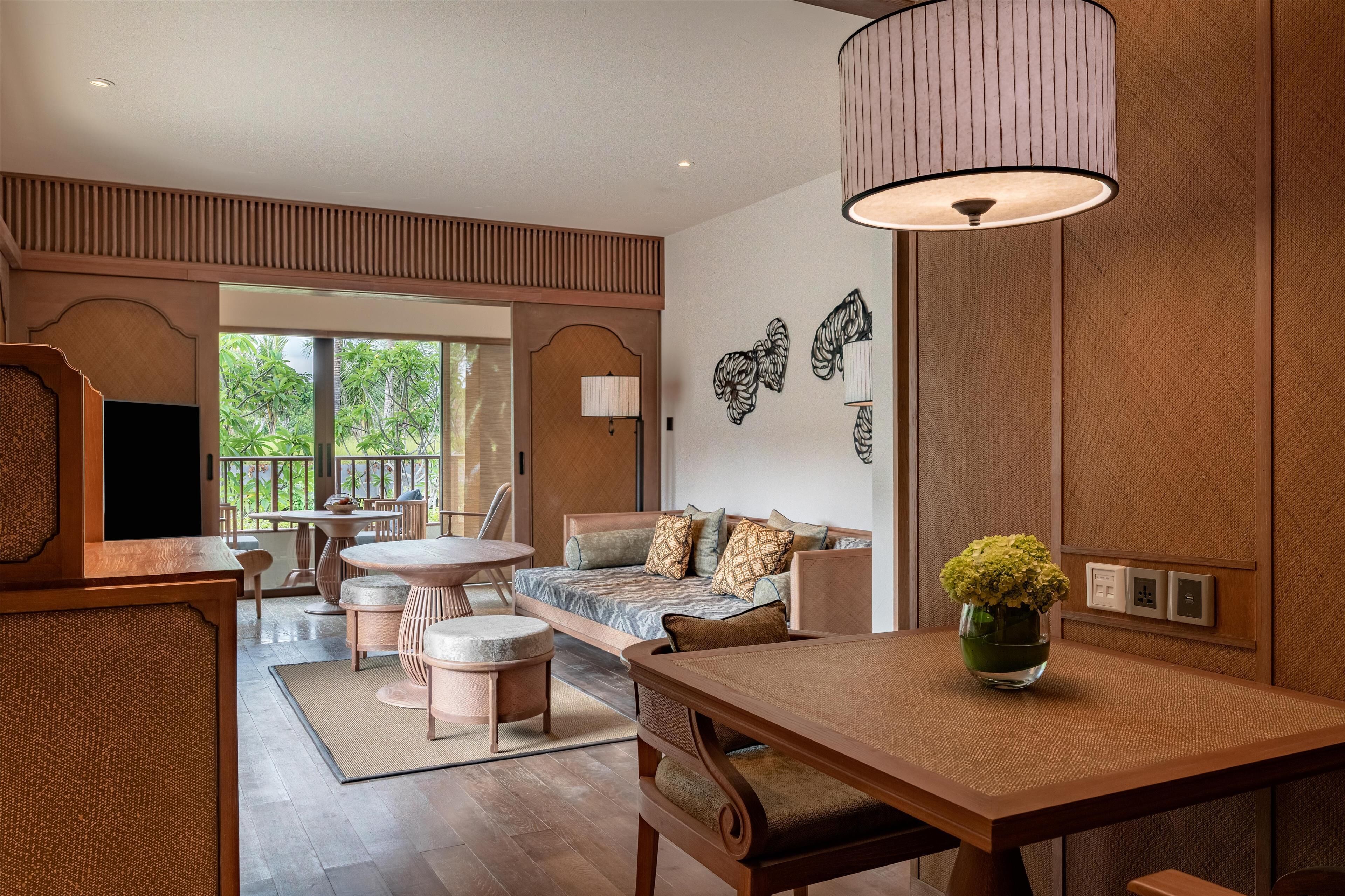 A modern hotel room with a wooden floor, a dining area with a table and chairs, a sofa with cushions, a large window with a view of greenery, and a wall-mounted lamp.