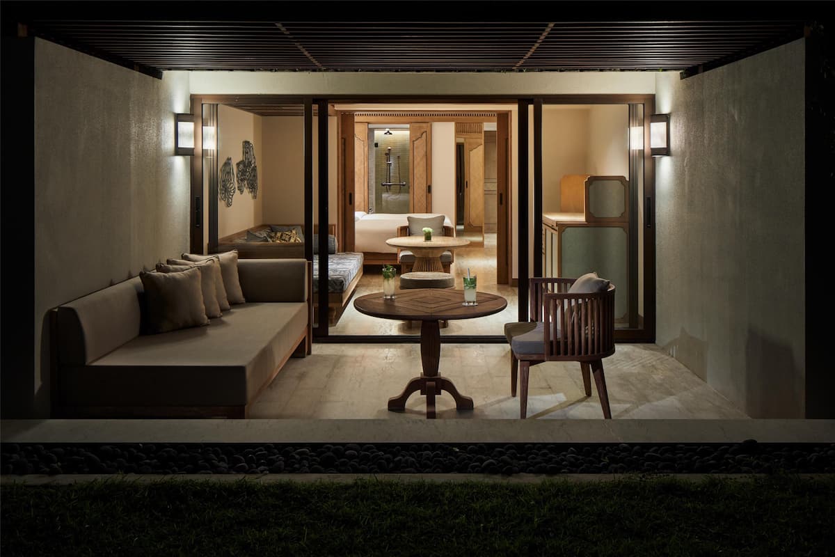 A modern hotel room with a sofa, a round table, and a chair. There is a bed with a nightstand and a lamp. The room has a large window and a view of the outside.