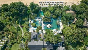An aerial view of a resort with a large swimming pool surrounded by lush greenery, a beach with umbrellas, and a boat in the water. - Hyatt Regency Bali (Denpasar)