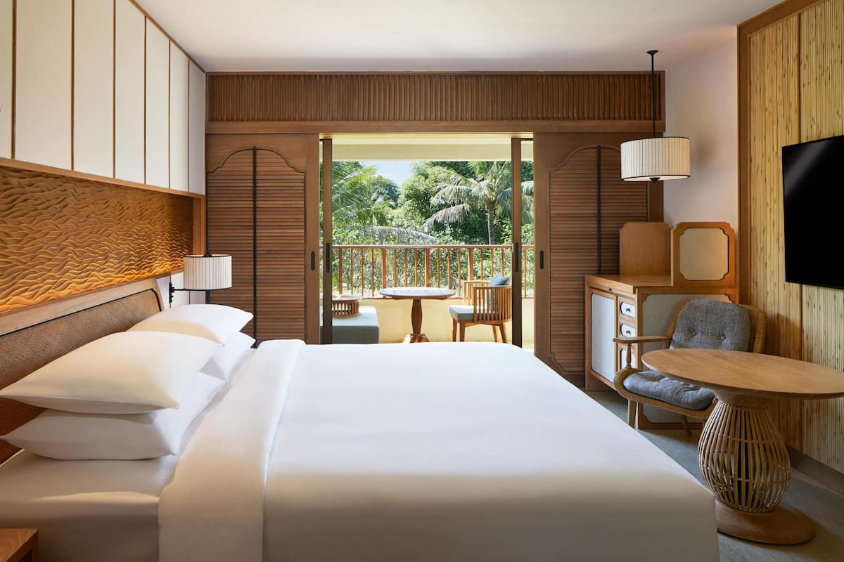 A hotel room with a bed, a small table, two chairs, a TV, and a balcony with a view of palm trees.