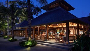 A resort with a covered outdoor dining area, palm trees, and a well-lit interior.