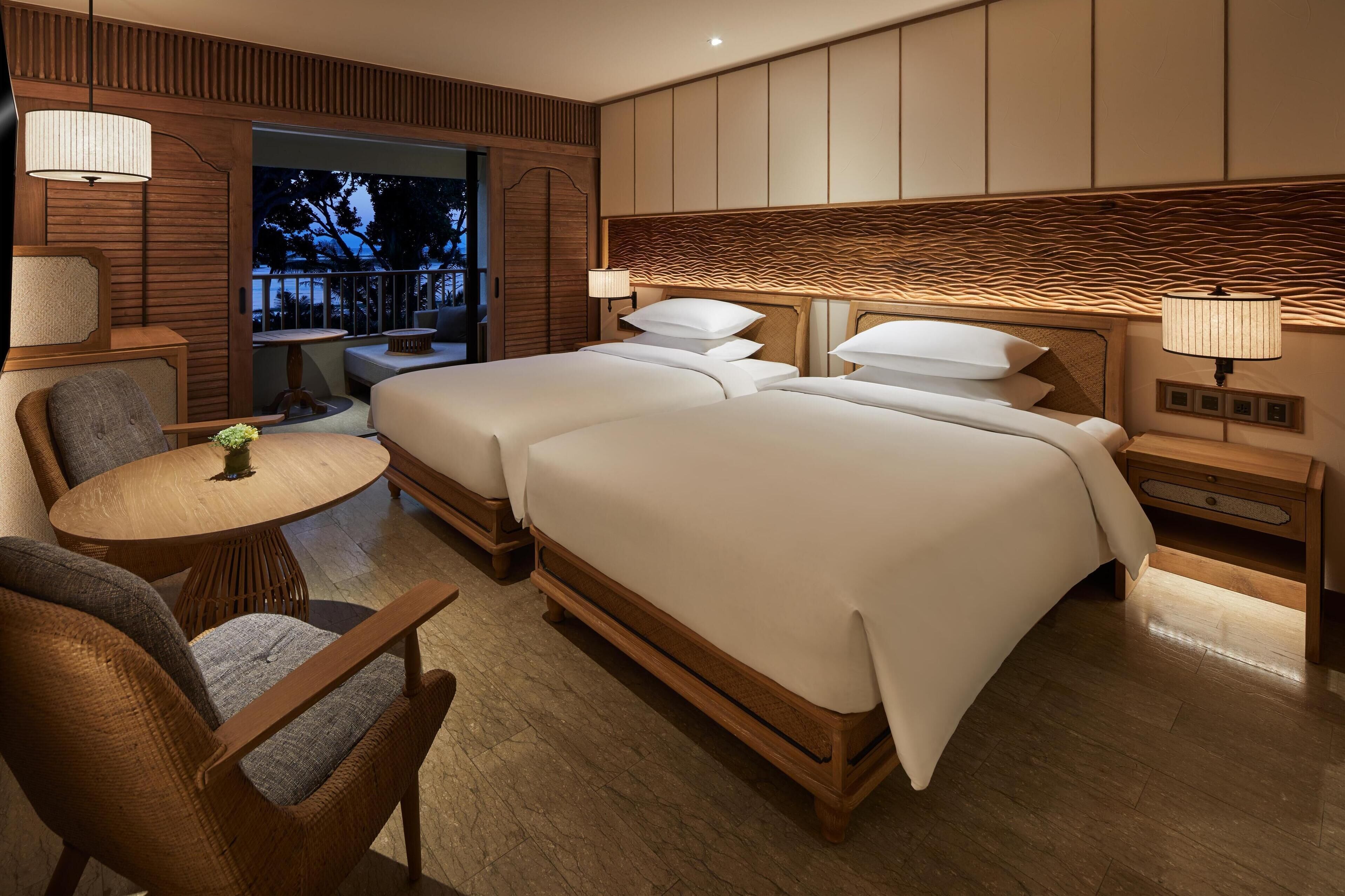 A hotel room with two beds, a sitting area with a table and chairs, and a balcony with a view of trees.