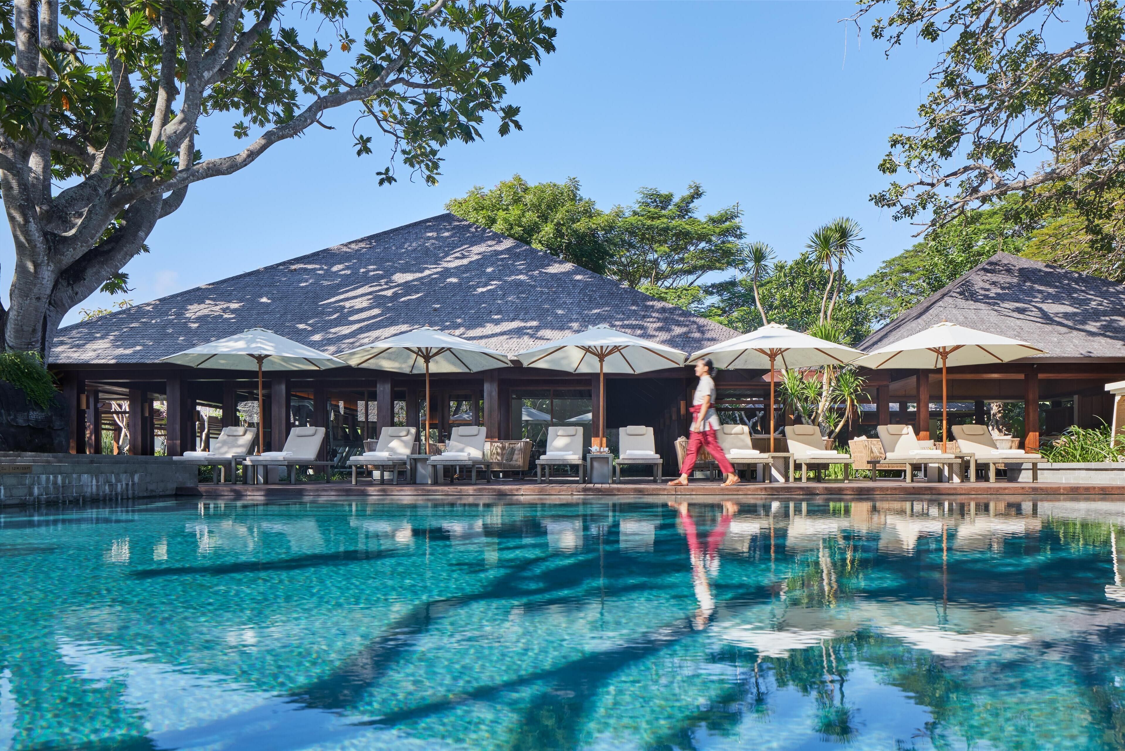 A pool area with lounge chairs and umbrellas, surrounded by trees and a building with a sloped roof.