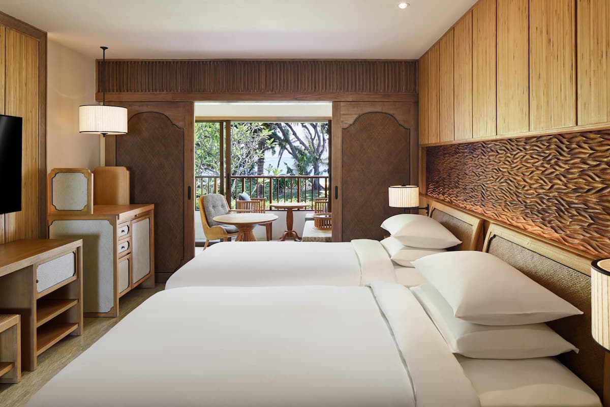 A hotel room with two beds, a desk, a chair, and a balcony with a view of palm trees.