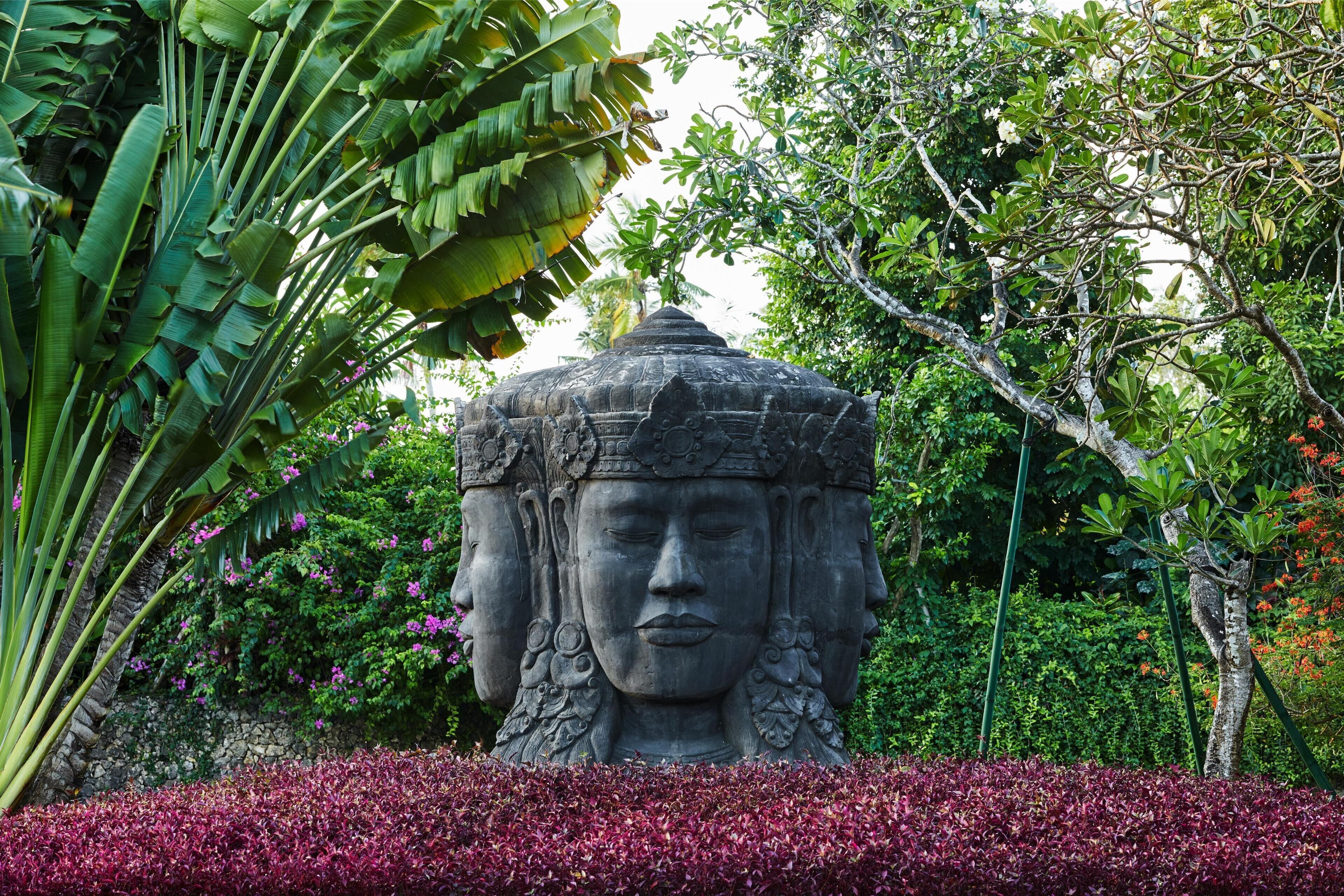A stone statue with intricate carvings, surrounded by lush greenery and flowering plants.