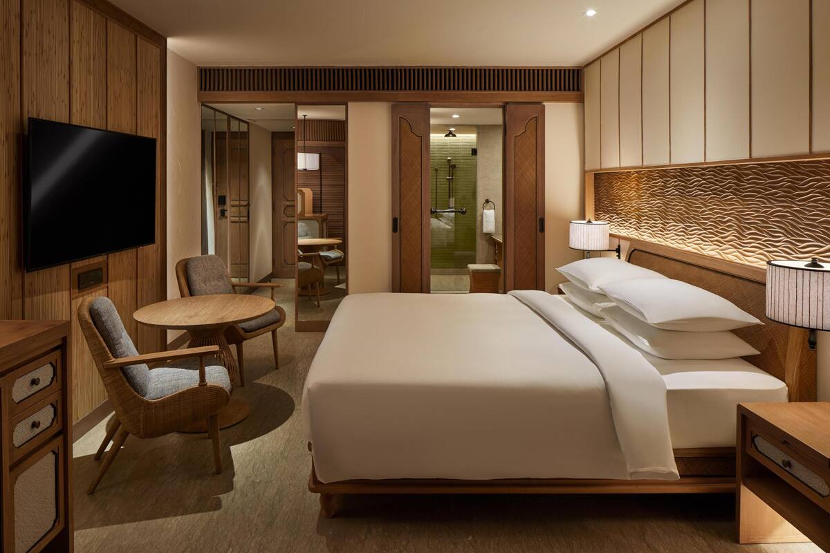 A hotel room with a large bed, a TV, a dining area with a table and chairs, and a bathroom visible through an open door.
