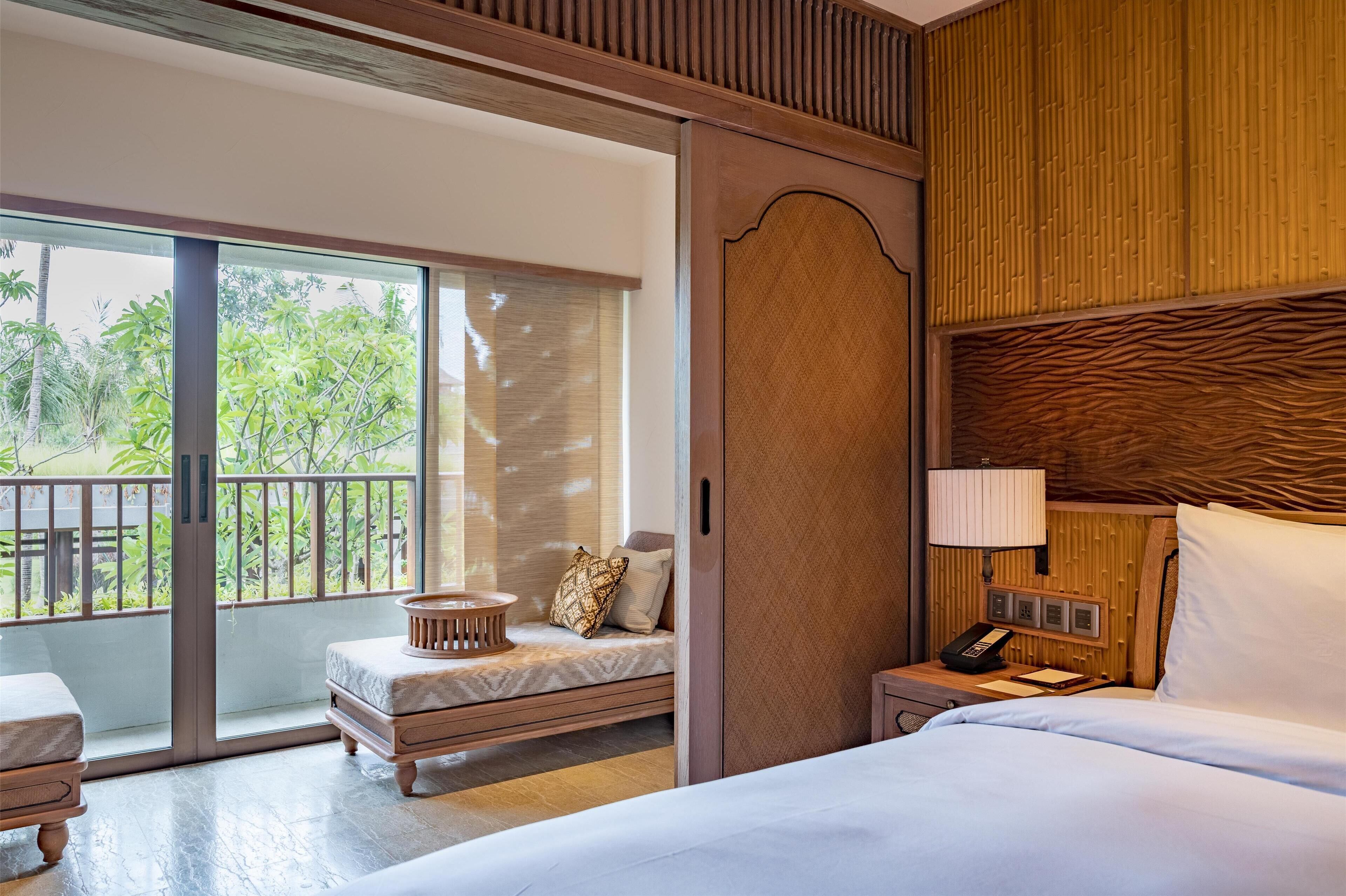 A hotel room with a bed, a bedside table with a lamp, a small seating area with a bench and cushions, and a sliding glass door leading to an outdoor balcony with greenery.