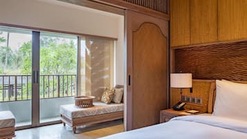 A hotel room with a bed, a bedside table with a lamp, a small seating area with a bench and cushions, and a sliding glass door leading to an outdoor balcony with greenery.