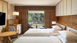 A hotel room with two beds, a TV, a desk, and a balcony with a view of trees.