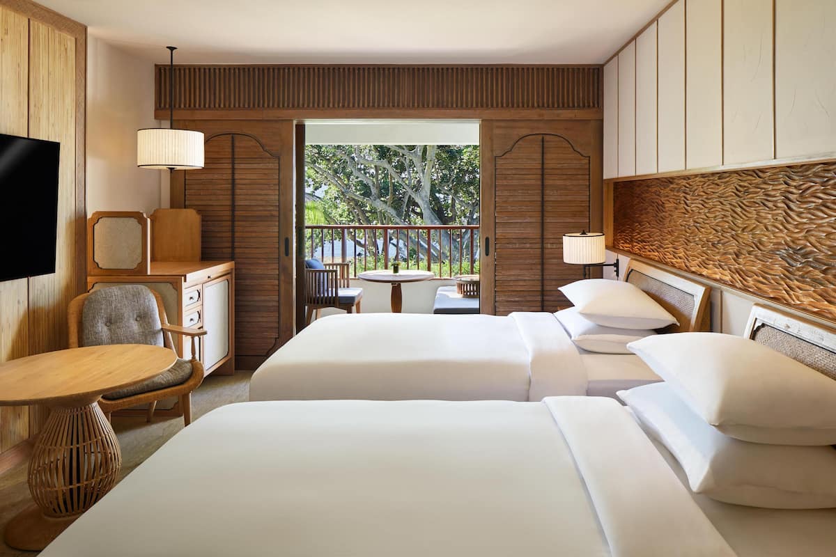 A hotel room with two beds, a TV, a desk, and a balcony with a view of trees.