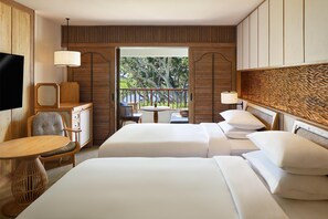 A hotel room with two beds, a TV, a desk, and a balcony with a view of trees.