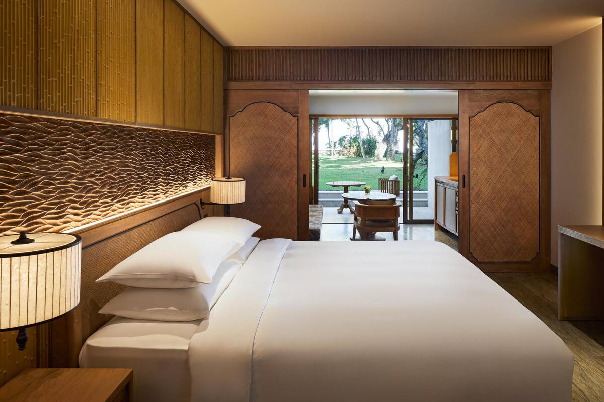 A hotel room with a large bed, a bedside table with a lamp, a view of the outdoor patio area with a table and chairs.