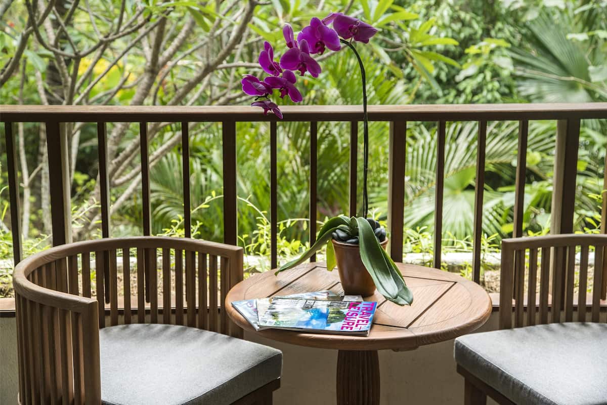 A wooden chair and table with a magazine, a potted plant, and purple orchids on a balcony with a view of greenery.