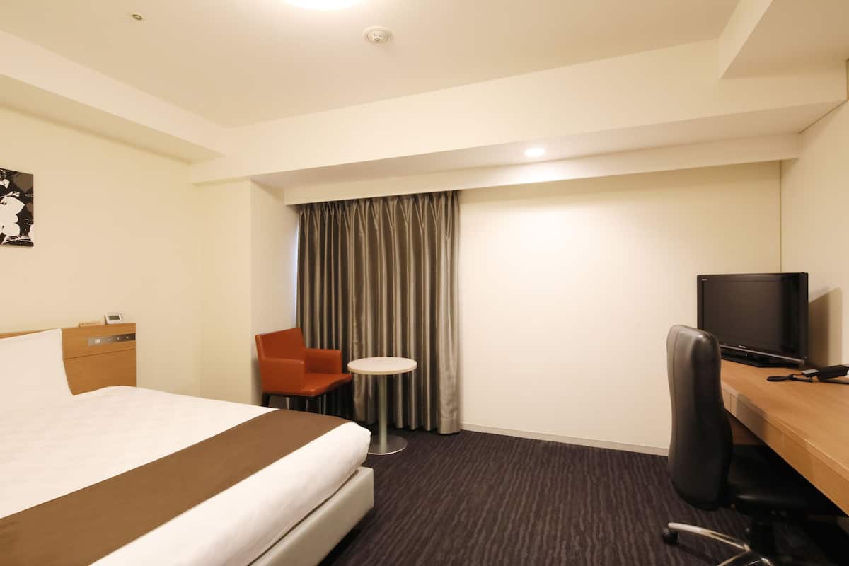 A hotel room with a bed, a desk with a computer, a chair, a small table, and a TV.