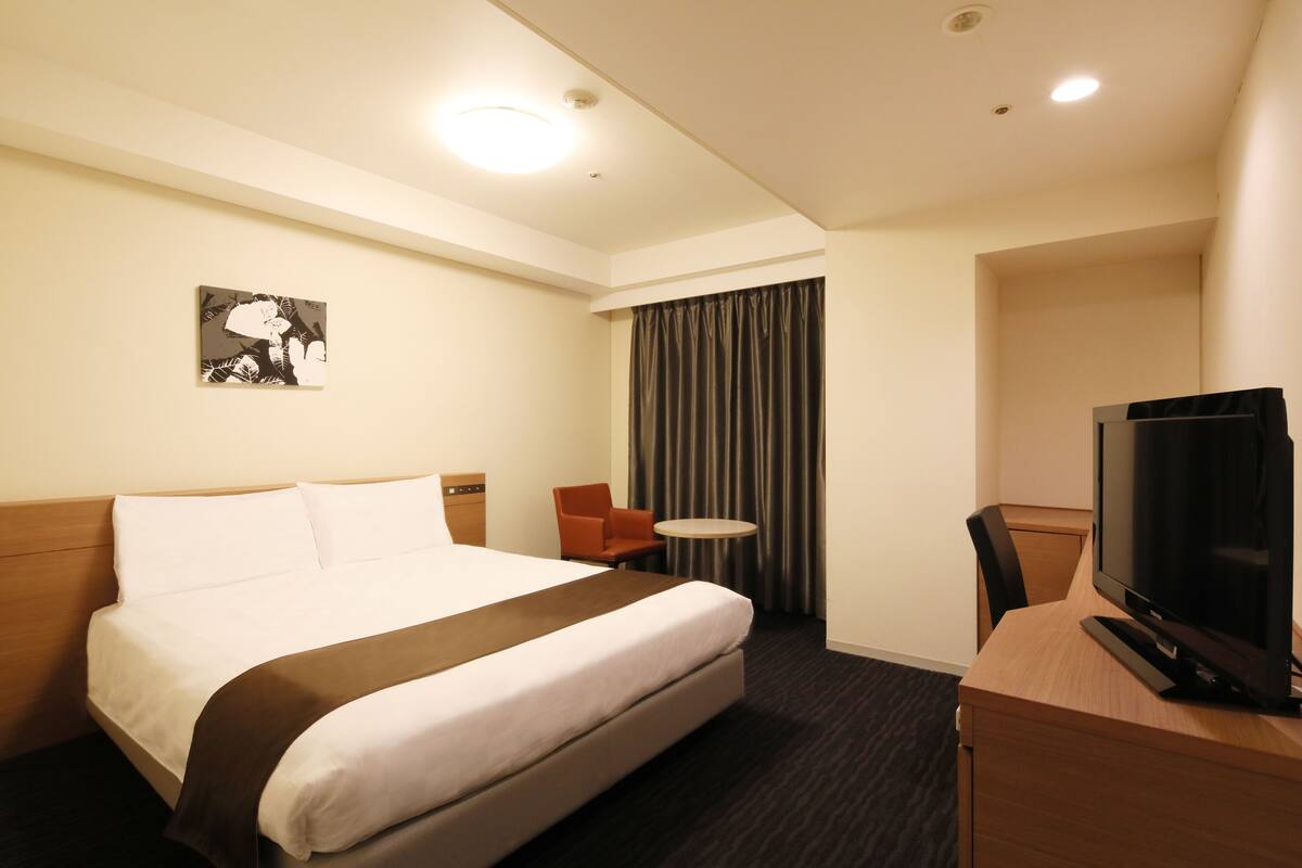 A hotel room with a large bed, a desk with a television, a chair, a small table, and a curtain.