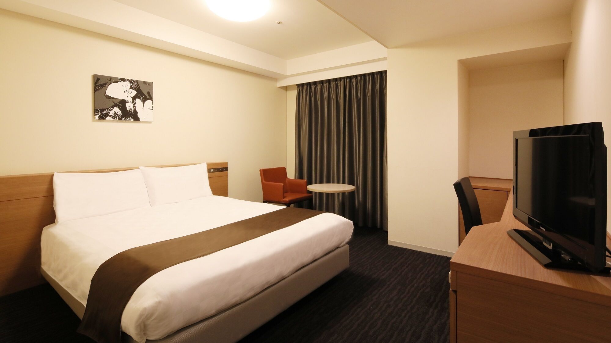 Habitación de hotel con cama, televisión, una silla, una mesita y un cuadro en la pared.