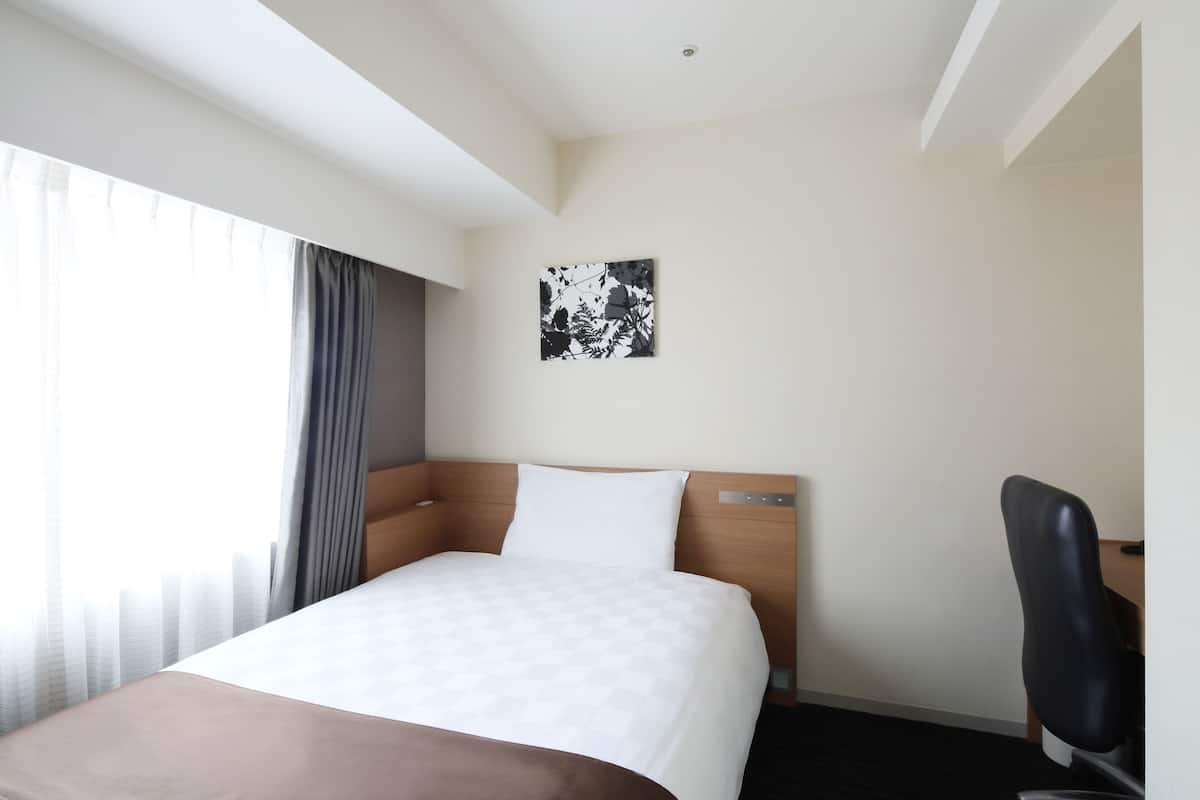 A hotel room with a bed, a desk, a chair, a window with curtains, and a wall-mounted artwork.