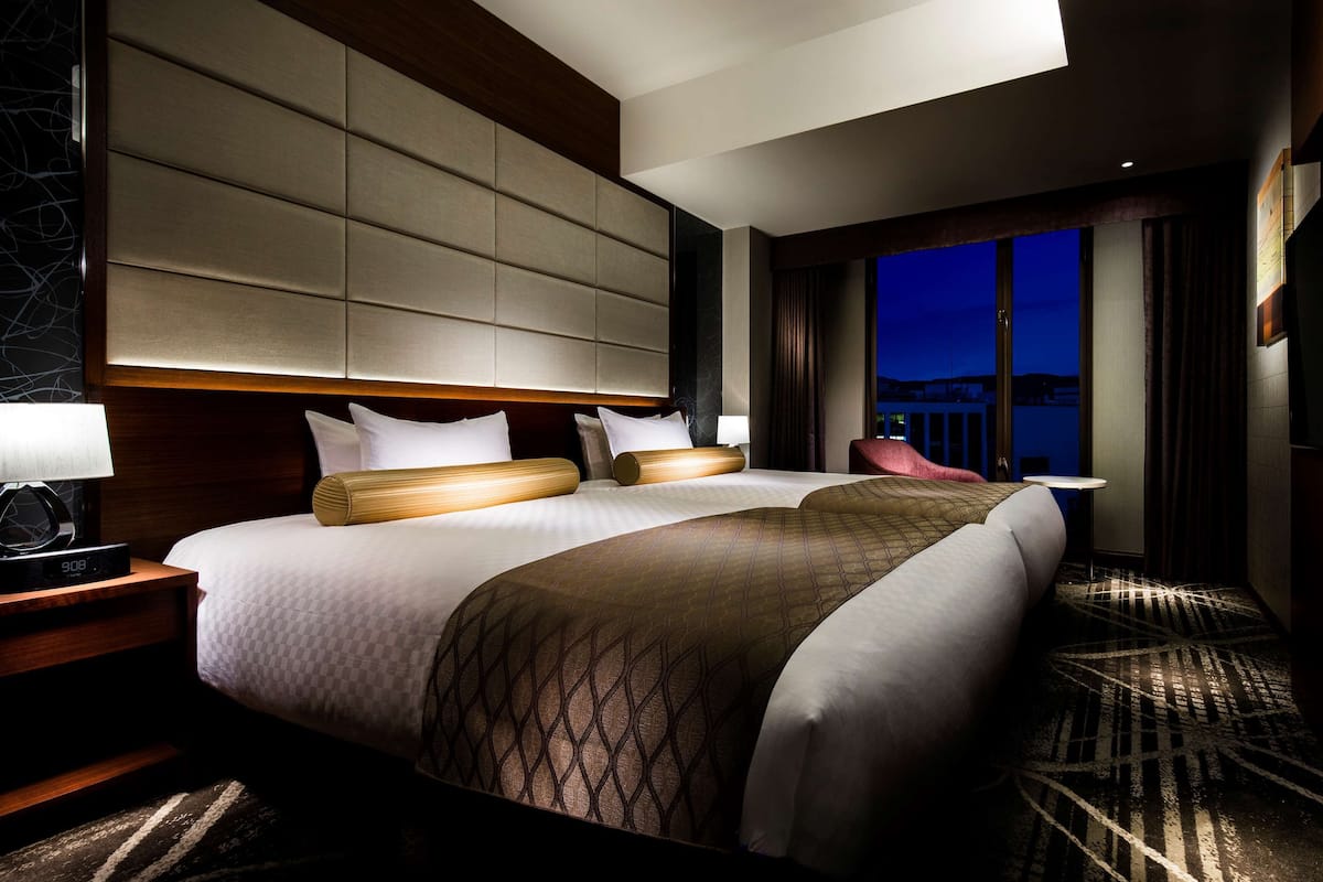 Une chambre d’hôtel avec un grand lit, deux tables de chevet, une commode, une lampe et une vue sur la ville la nuit.