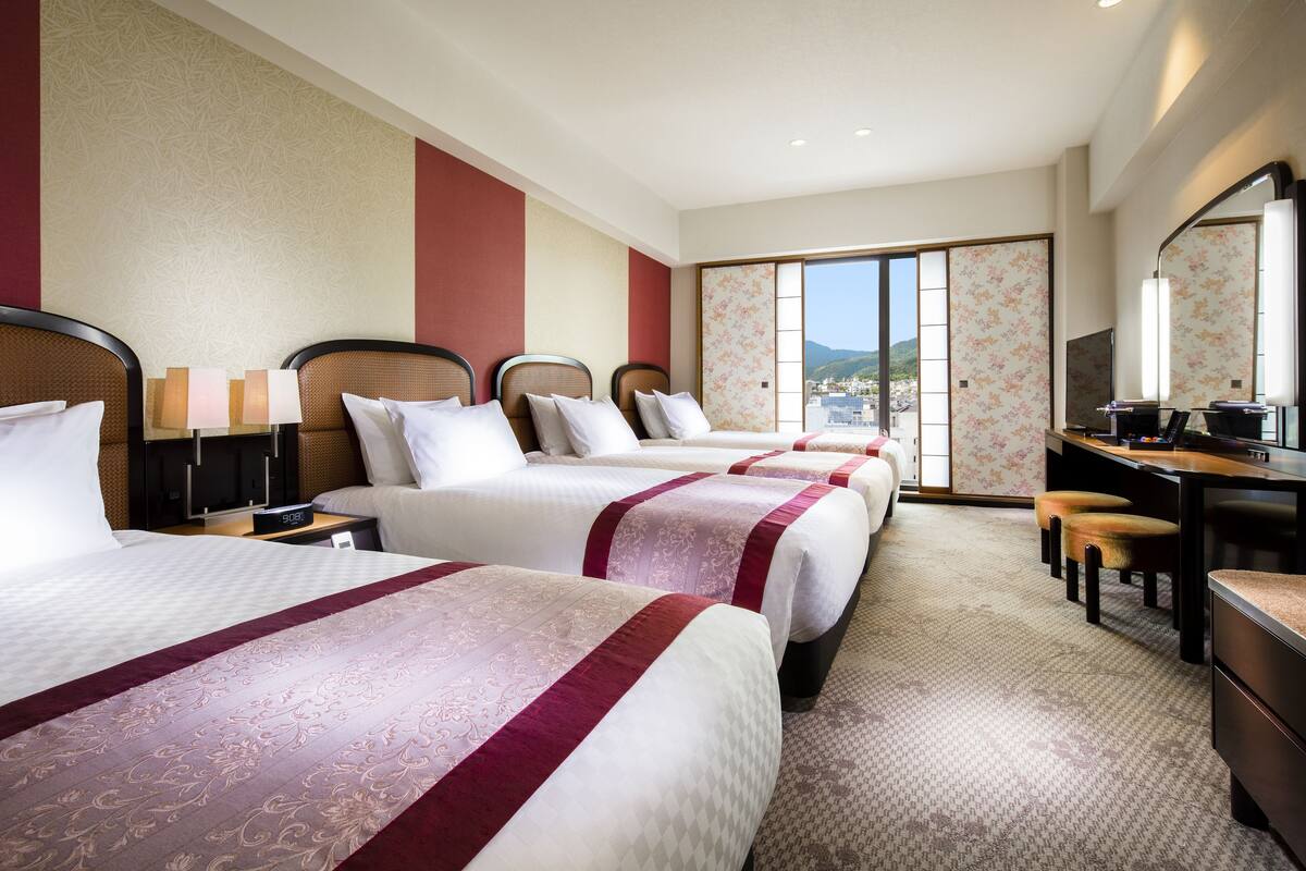 Une chambre d’hôtel avec trois lits, un bureau et une grande fenêtre donnant sur un beau paysage.