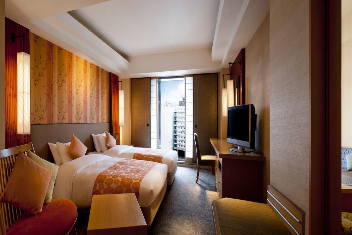 Une chambre d’hôtel avec deux lits, un bureau, une télévision et une vue sur un immeuble par la fenêtre.