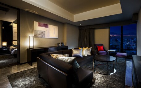 A modern hotel room with a dark leather sofa, a small round table, a flat-screen TV, and a city view through large windows.