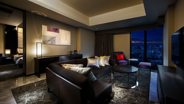 Una habitación de hotel moderna con un sofá de cuero oscuro, una pequeña mesa redonda, un televisor de pantalla plana y vistas a la ciudad a través de amplias ventanas.