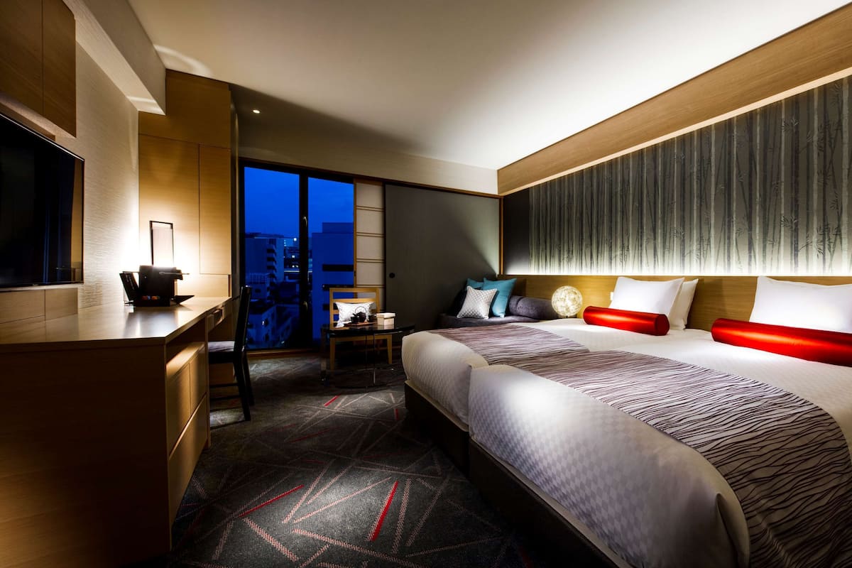 Une chambre d’hôtel moderne dotée d’un grand lit, d’un bureau avec une machine à café, d’une chaise, d’une vue sur la ville au crépuscule et d’une grande fenêtre.