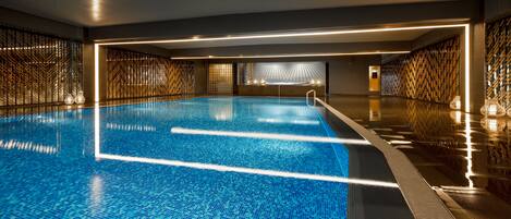 Een groot binnenzwembad met helderblauw water, omringd door muren met een geometrisch patroon en warme verlichting.