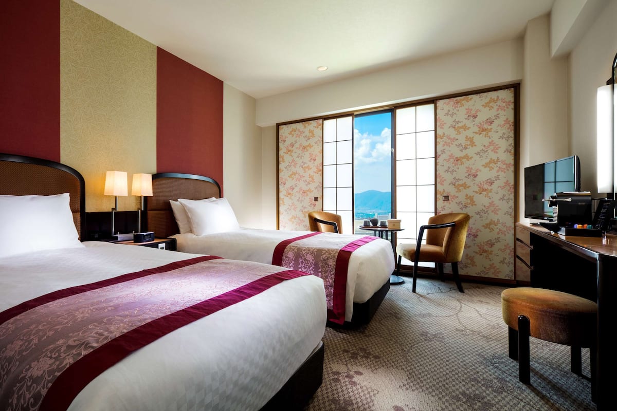 Une chambre d’hôtel avec deux lits, un bureau équipé d’un ordinateur, une chaise et une vue sur les montagnes grâce à de grandes fenêtres.