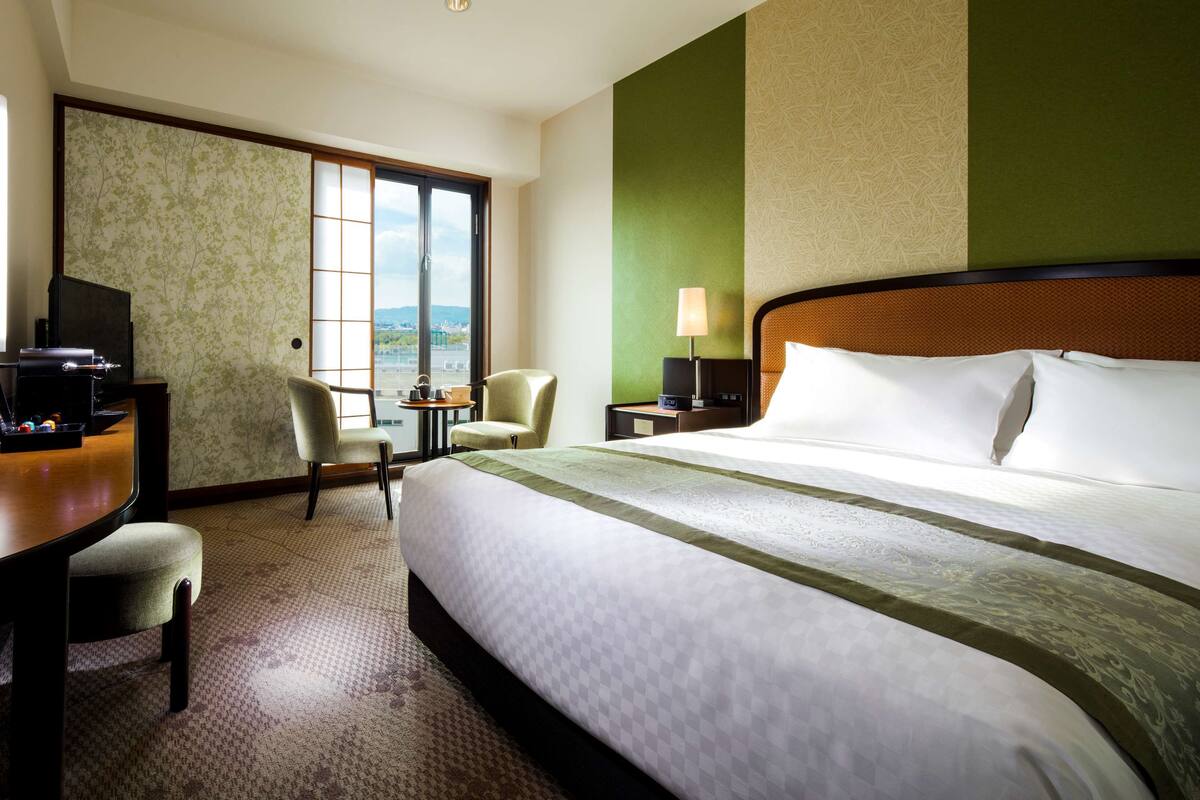 Une chambre d’hôtel avec un grand lit, deux fauteuils, un bureau avec une télévision et une vue sur l’extérieur.