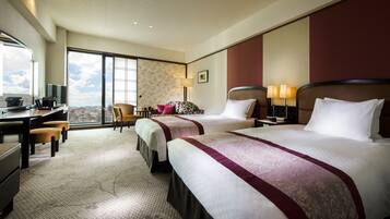 A hotel room with two beds, a desk, a chair, and a view of the city.