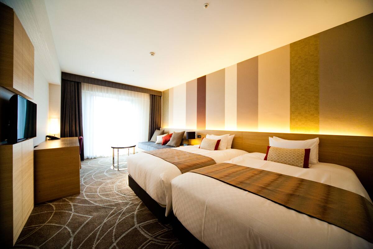 Une chambre d’hôtel avec deux lits, un bureau, une télévision et une grande fenêtre avec des rideaux.