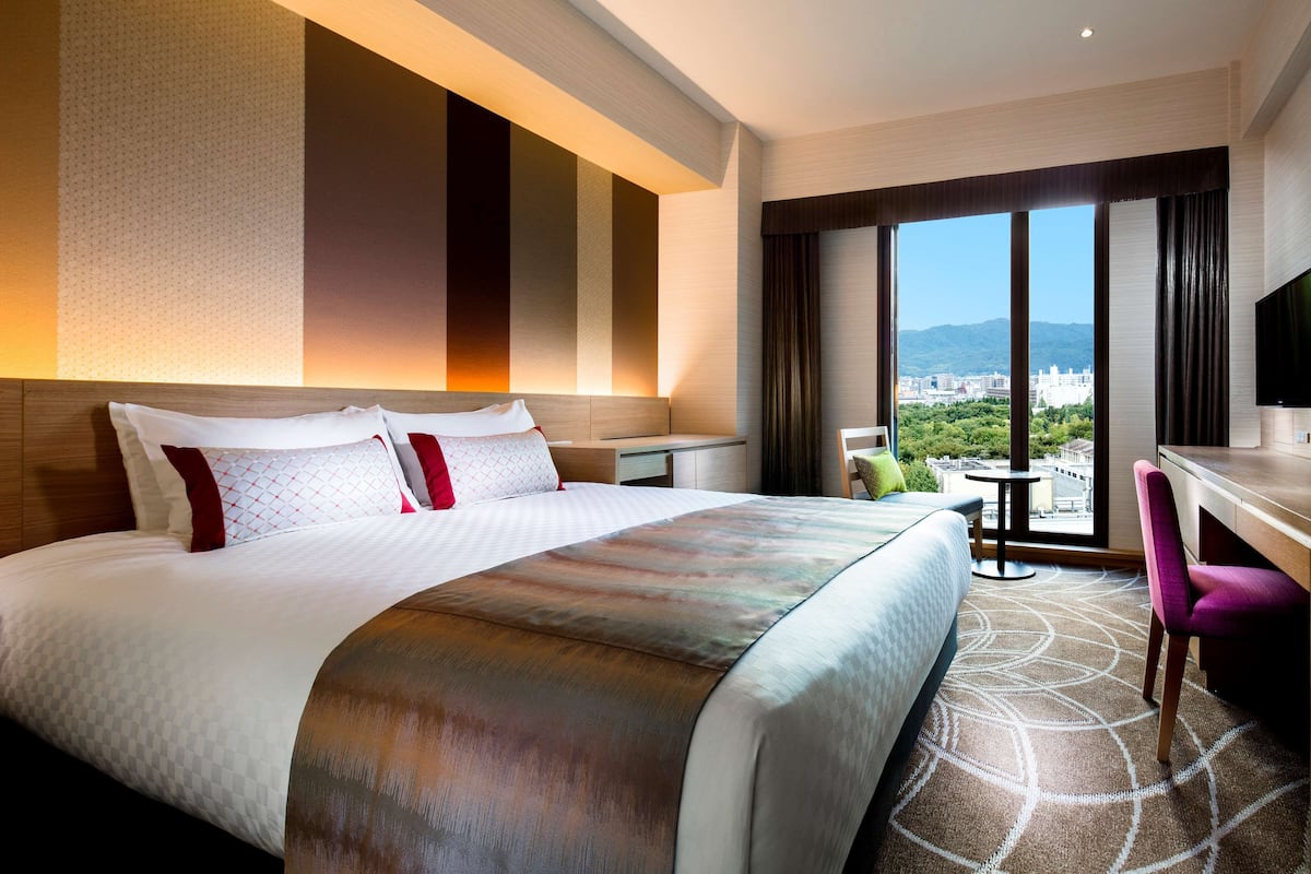 Une chambre d’hôtel avec un grand lit, un bureau, une chaise et une vue sur le paysage urbain, visible à travers une grande fenêtre.