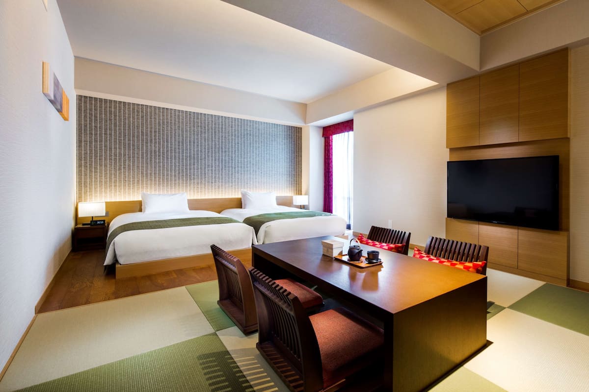 Une chambre d’hôtel avec deux lits, une table à manger avec des chaises, un téléviseur et une grande fenêtre avec des rideaux.