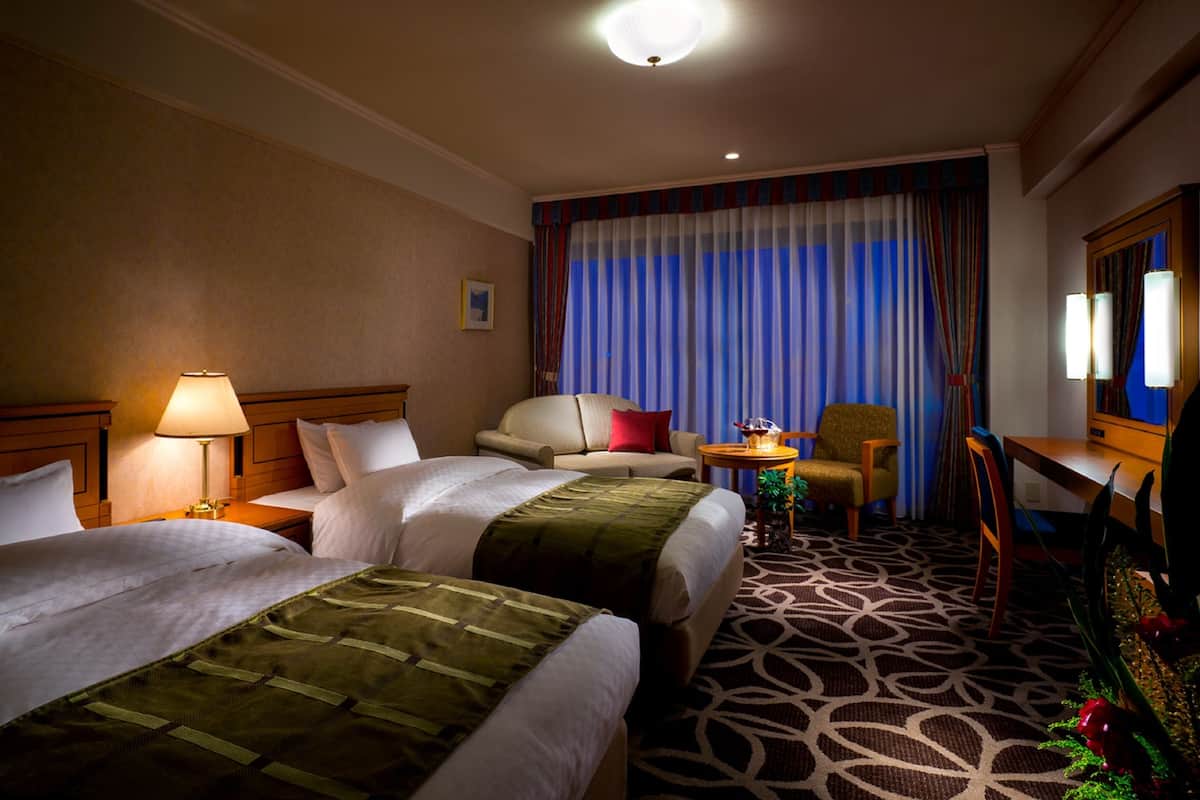 Une chambre d’hôtel avec deux lits, un bureau, une chaise, une lampe et une grande fenêtre avec des rideaux.