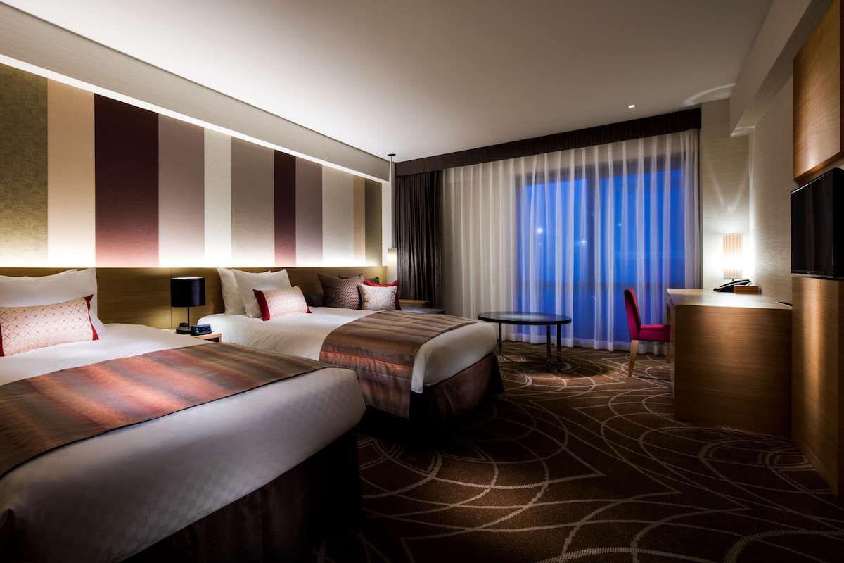 Une chambre d’hôtel avec deux lits, un bureau, une chaise, une télévision et une grande fenêtre avec des rideaux.