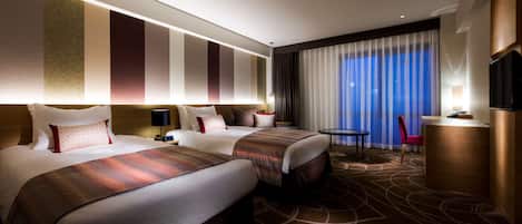 A hotel room with two beds, a desk, a chair, a TV, and a large window with curtains.
