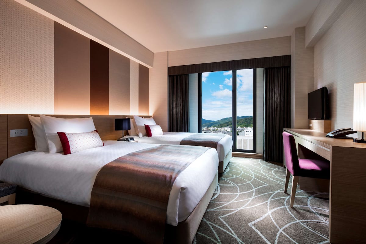 Une chambre d’hôtel avec deux lits, un bureau, une chaise et une grande fenêtre donnant sur la ville.