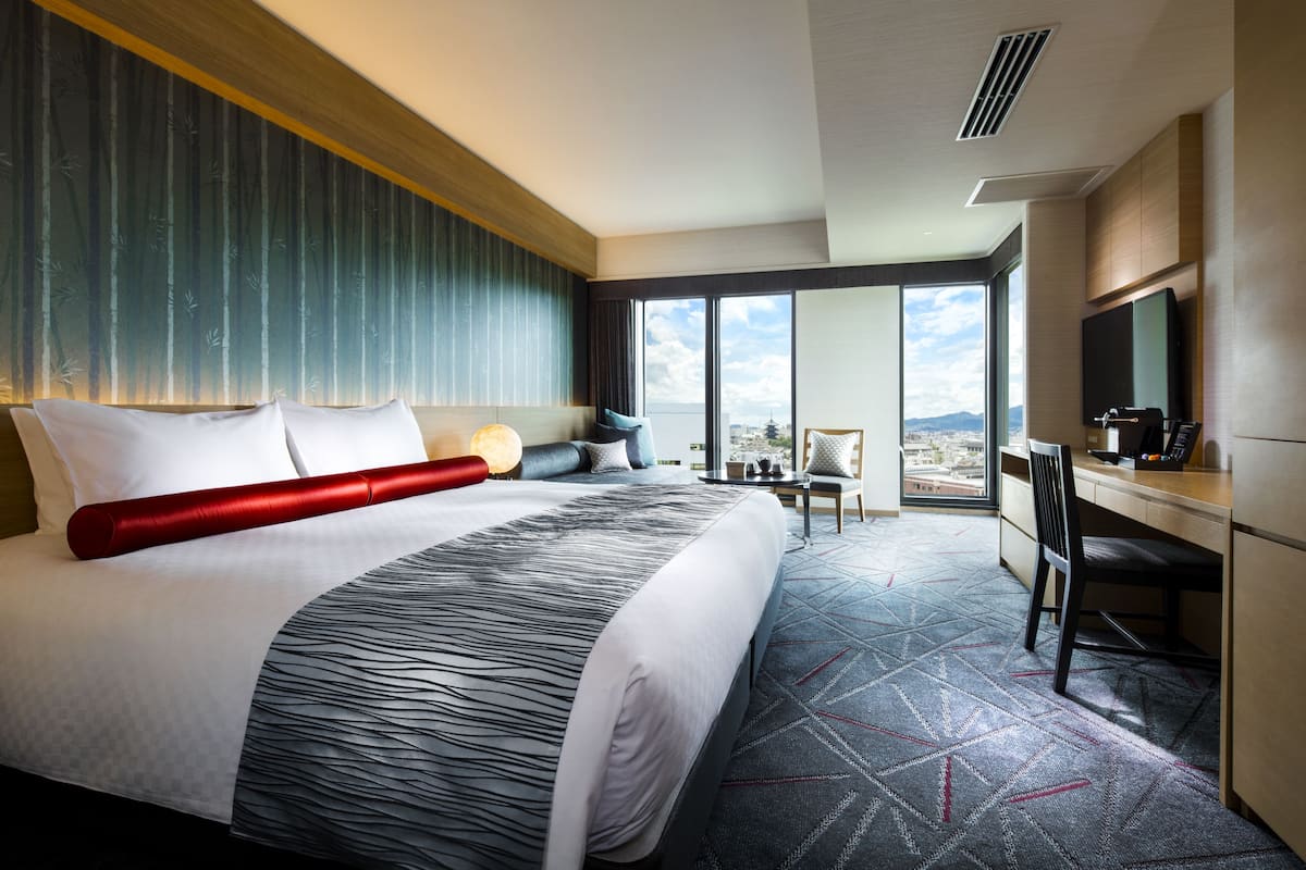 Une chambre d’hôtel moderne dotée d’un grand lit, d’un bureau avec un ordinateur, d’une télévision et offrant une vue sur le paysage urbain.