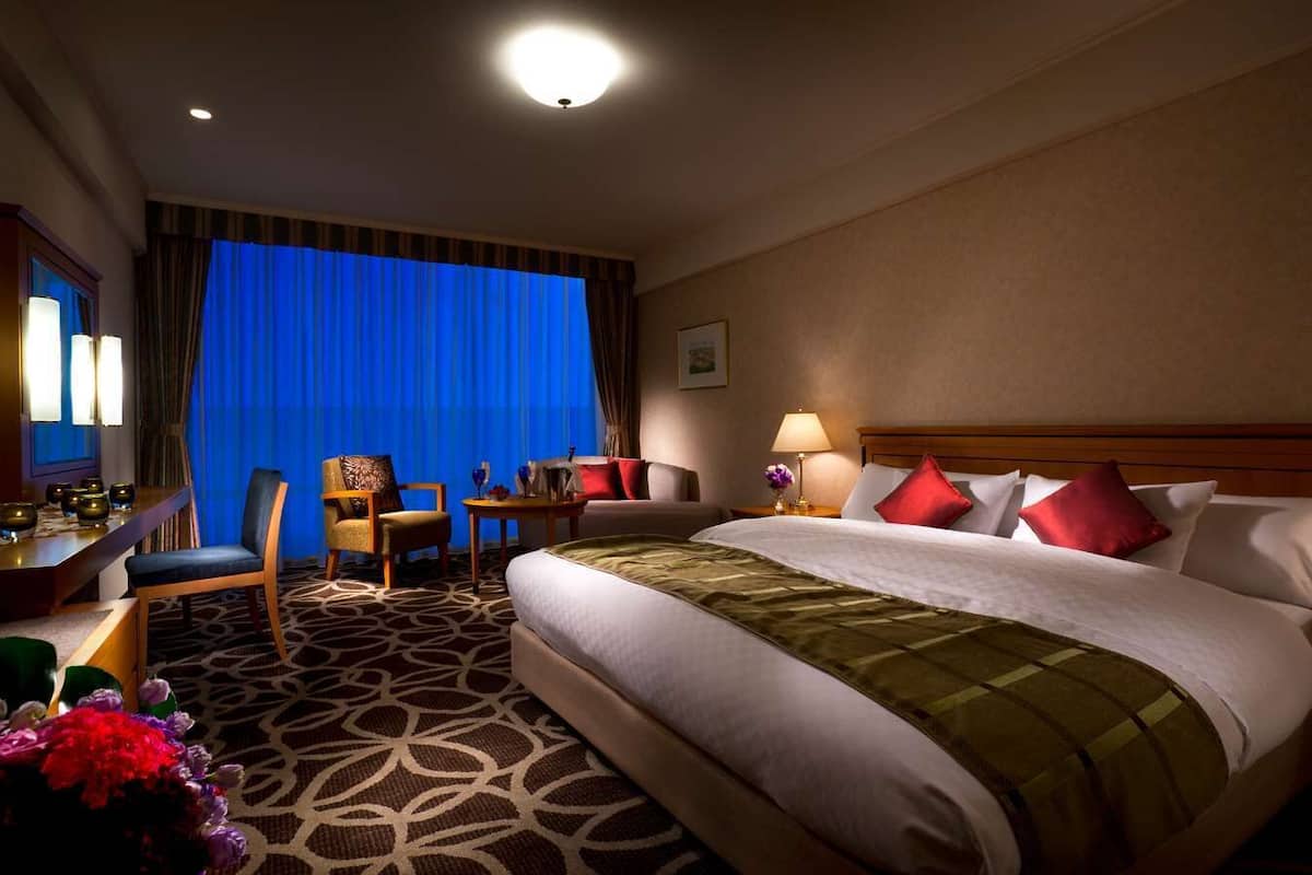 Une chambre d’hôtel avec un grand lit, un bureau avec une lampe, une chaise et une vue sur la ville par la fenêtre.
