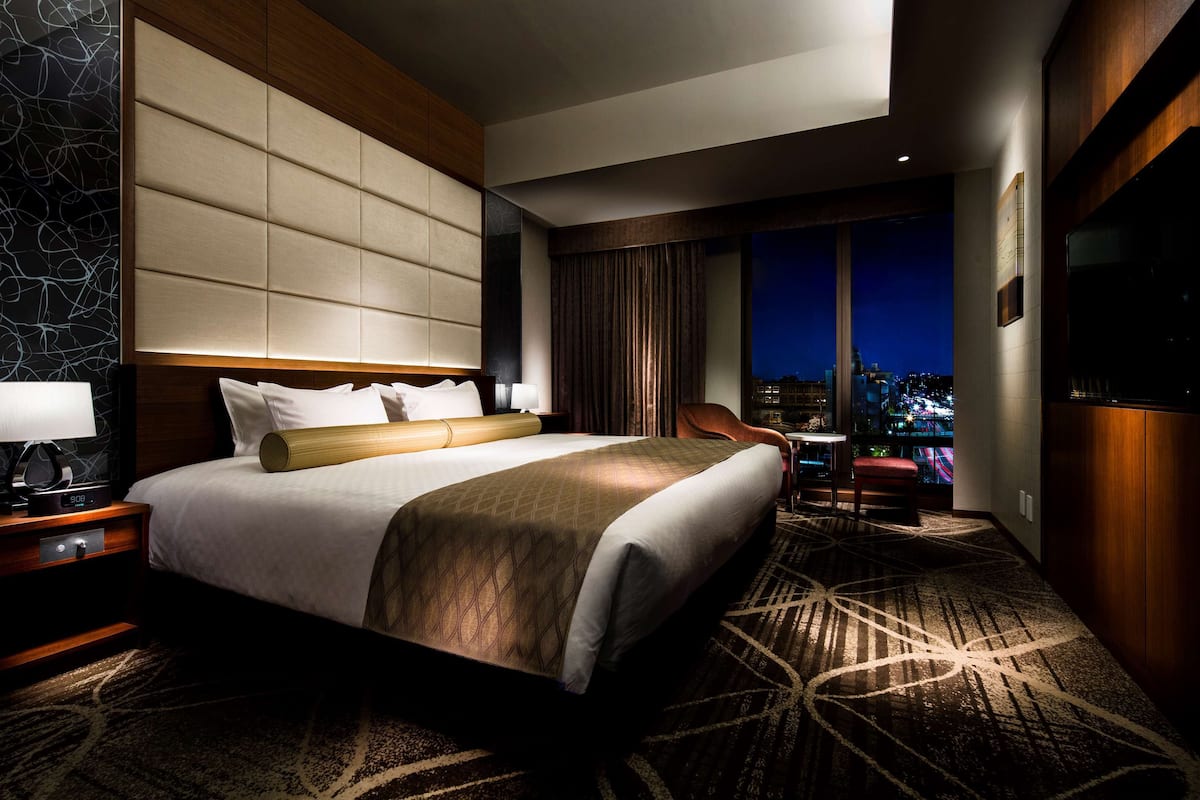 Une chambre d’hôtel avec un grand lit, une table de chevet, un bureau, une chaise et une vue sur la ville la nuit.