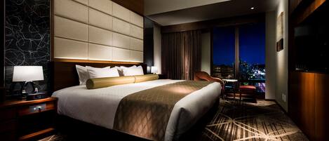 Un cuarto de hotel con una cama grande, una mesita de noche, un escritorio, una silla y vistas de la ciudad iluminada.
