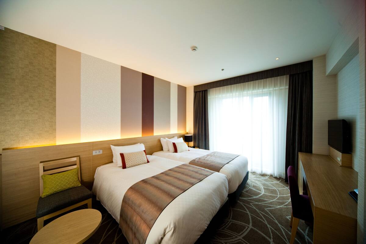 Une chambre d’hôtel avec deux lits, un bureau, une chaise, une télévision et une fenêtre avec des rideaux.