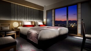 Una habitación de hotel moderna con una cama grande, una mesita de noche, una silla y vistas a la ciudad al anochecer.