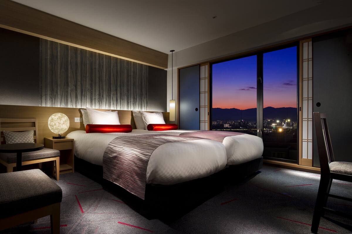 Une chambre d’hôtel moderne avec un grand lit, une table de chevet, une chaise et une vue sur la ville au crépuscule.