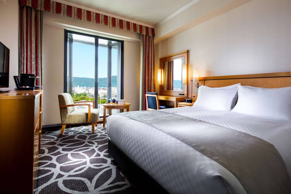 Une chambre d’hôtel dotée d’un grand lit, d’un bureau, d’une chaise, d’une télévision et offrant une vue sur la ville grâce à de grandes fenêtres.