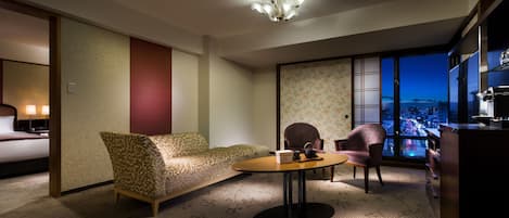 Habitación de hotel con sofá, sillas, una mesita, cama y vistas a la ciudad a través de grandes ventanales.