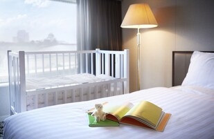 A hotel room with a crib, a bed with a book and a teddy bear, a lamp, and a view of the city.
