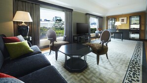 A modern hotel room with a sofa, chairs, a coffee table, a TV, and large windows offering a city view.