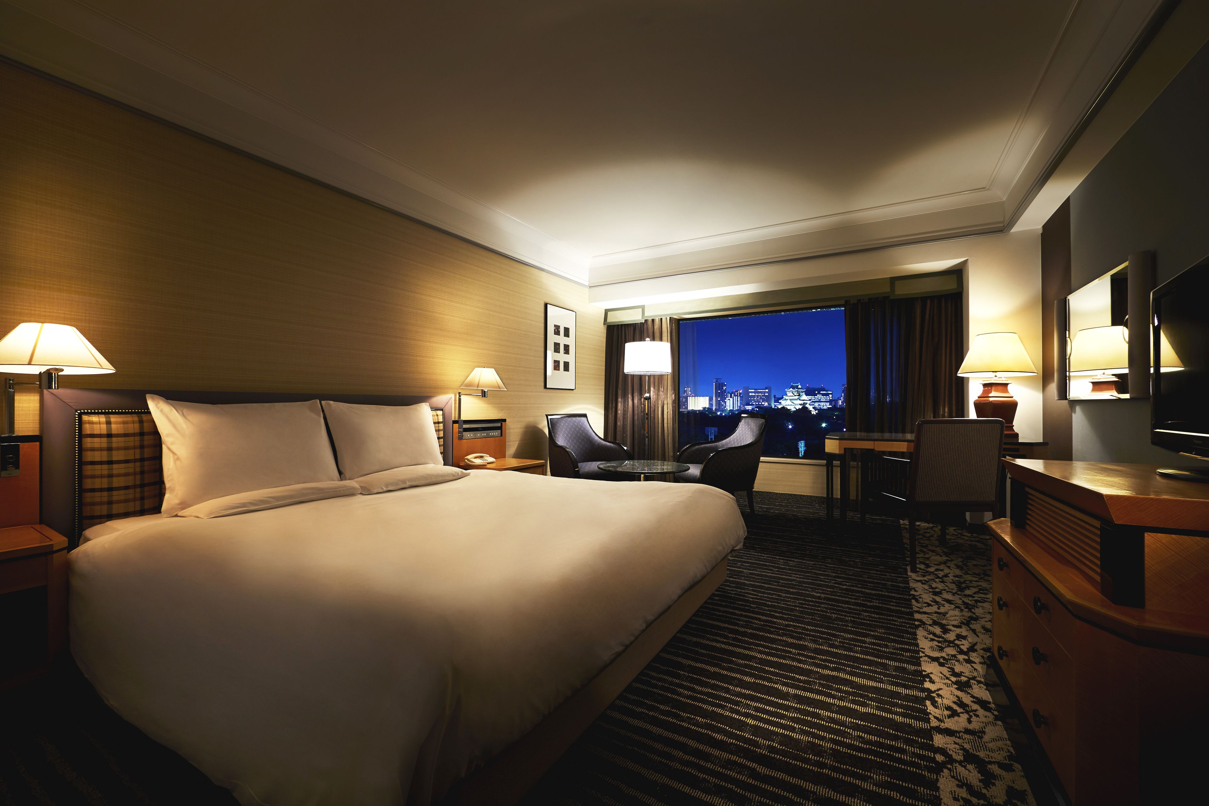 A hotel room with a large bed, two bedside tables with lamps, a desk with a chair, and a view of the city at night.